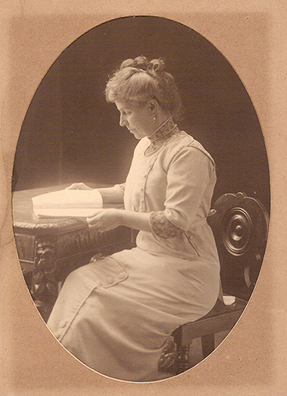 A sepia toned historical image of a woman sitting at a carved wooden table, reading a document. She is wearing a high necked dress, with three quarter length sleeves, Her hair is coiled off her face, and she is concentrating.