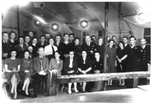 A black and white photo of members of the Polish Navy. A large group of men and women stand in a building with a curved / domed ceiling. There are a few bright lights hanging on cables, and a wooden post and low fence in the foreground of the image. Most of the men are in naval uniform. Most of the women are in smart dresses. 