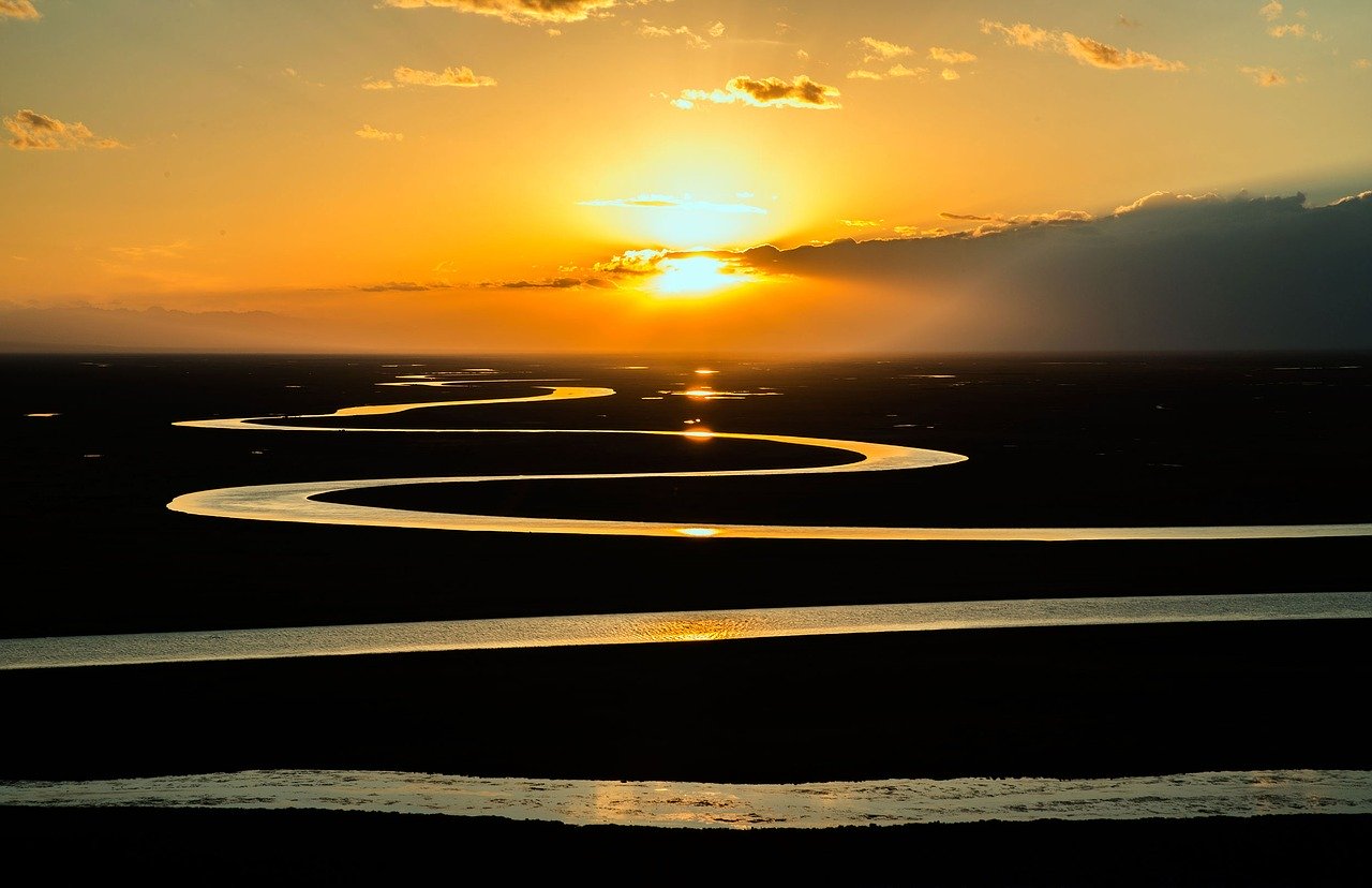 A photograph of a sunrise. The sky is filled with orangey tones, and the sun is partly obscured by fine cloud. In the foreground, a river snakes away in a repeating 'S' shape: wider at the bottom and growing narrower as it moves toward the horizon at the top of the photograph.