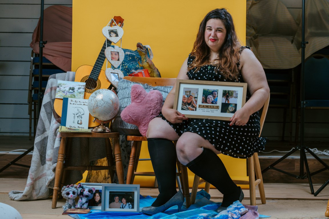 Ingrid, a young woman of Spanish heritage, sits on a chair holding a framed series of photos of her family. It says 'family' in the centre on Scrabble tiles. She is pictured against a yellow backdrop and surrounded by meaningful objects, including: photos, books, a cushion, toys and a guitar (photo: Dom Moore)